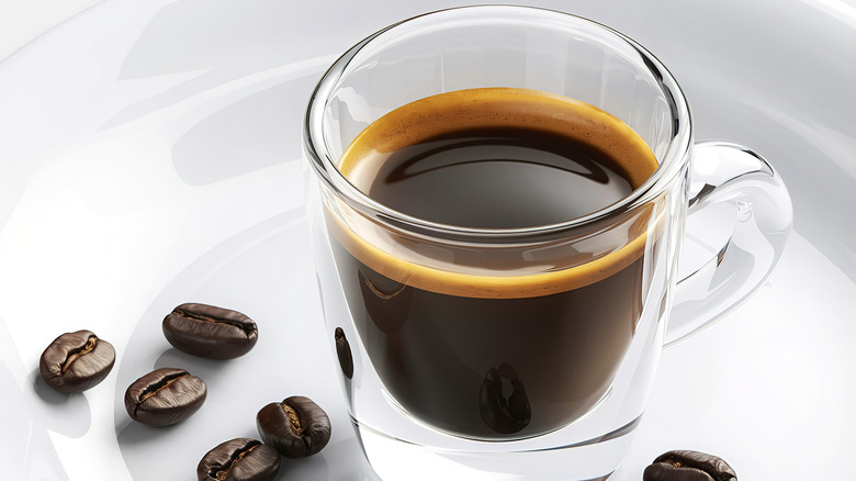 A closeup view of an espresso shot glass filled with espresso with coffee beans around on a white texturized background