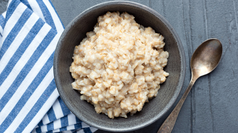 A bowl of cooked steel-cut oatmeal on a gray surface near a blue-and-white striped cloth and a metal spoon