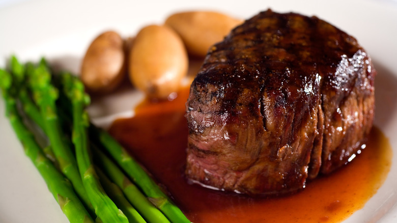 A filet mignon steak on a plate with sauce, asparagus, and potatoes