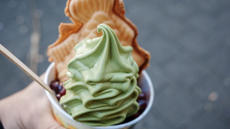 A hand holds a cup of matcha ice cream with sweet red beans and a Japanese fish cake