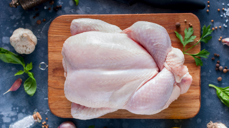 A whole raw chicken sits on a wooden cutting board surrounded by herbs and aromatics