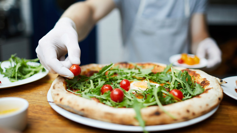 Hands topping a pizza with arugula and tomato
