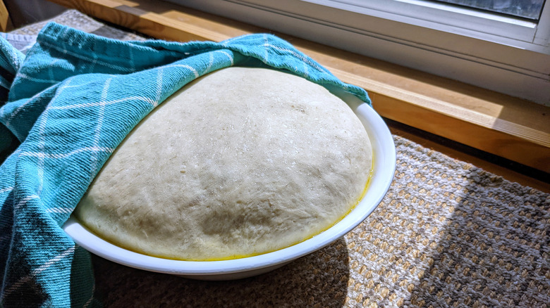 A bowl of bread dough covered slightly by a blue towel sits in the sun