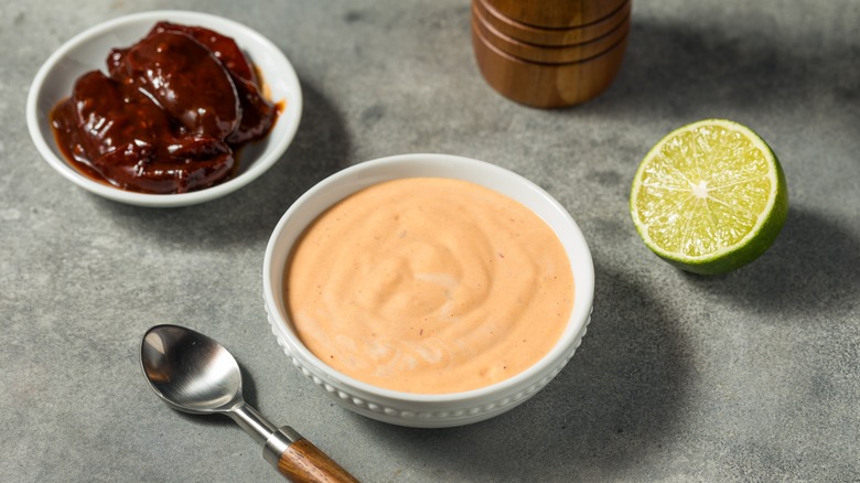 Small white bowl of chipotle lime mayo next to chipotle peppers and lime half