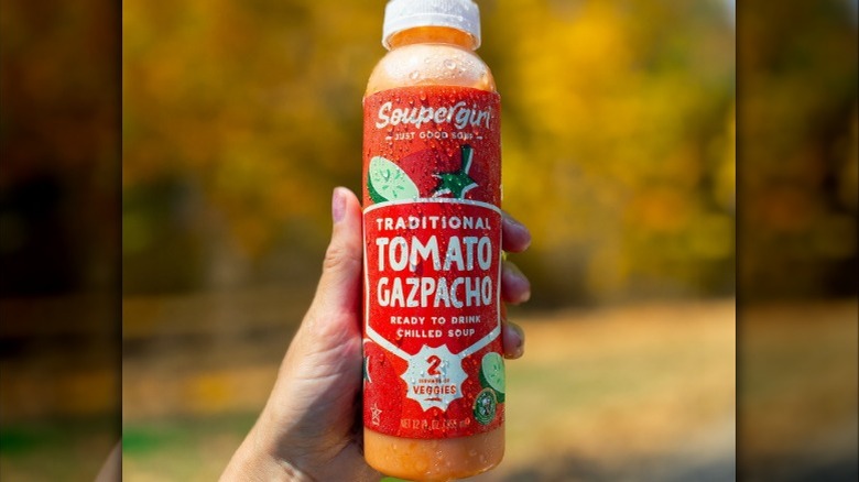 A hand holds a bottle of tomato gazpacho with an autumnal background