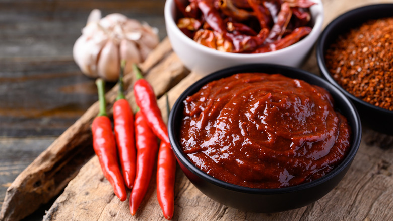 Small black bowl of red gochujang sauce on a wooden surface with another black bowl next to it with red paper flakes and a white bowl in the background with whole dried red peppers, as well as a head of garlic in the back and five thin fresh red peppers not to the front bowl