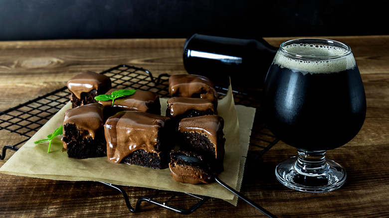 Rounded glass of dark beer on a wooden surface next to several small chocolate frosted brownies on parchment paper on top of a black rack