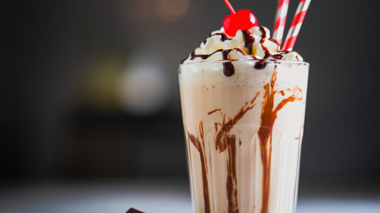 A chocolate milkshake in a glass cup with whipped cream and chocolate sauce and a cherry on top