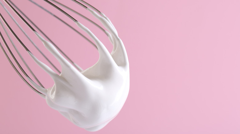 Whipped cream sticks to a wire whisk in front of a pink background.