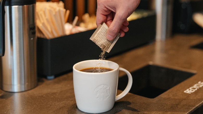 A Starbucks customer sprinkles a packet of sugar into their coffee held in a white ceramic mug