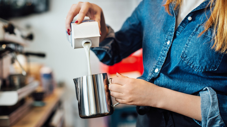 A barista wearing a blue denim shirt pours milk into a silver canister