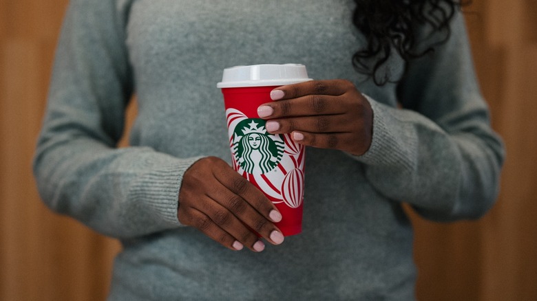 A woman wearing a grey sweater holds a red reusable Starbucks cup