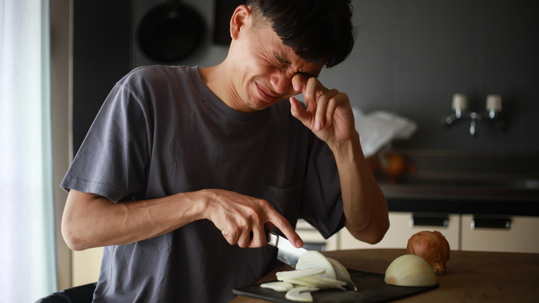 Crying man cutting onions with knife, wiping tears from eyes