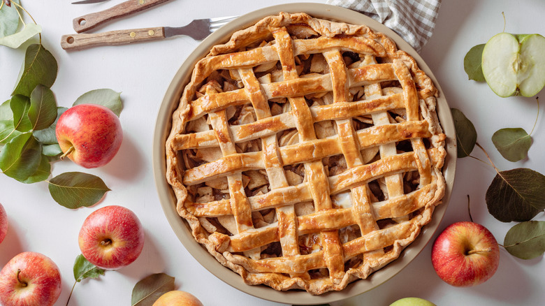 An apple pie with pretty lattice is surrounded by fresh apples