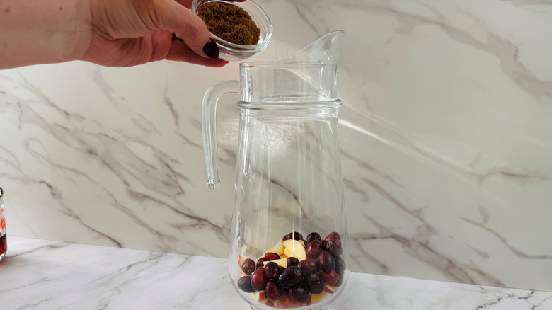 Hand adding brown sugar to a glass jug with fruit