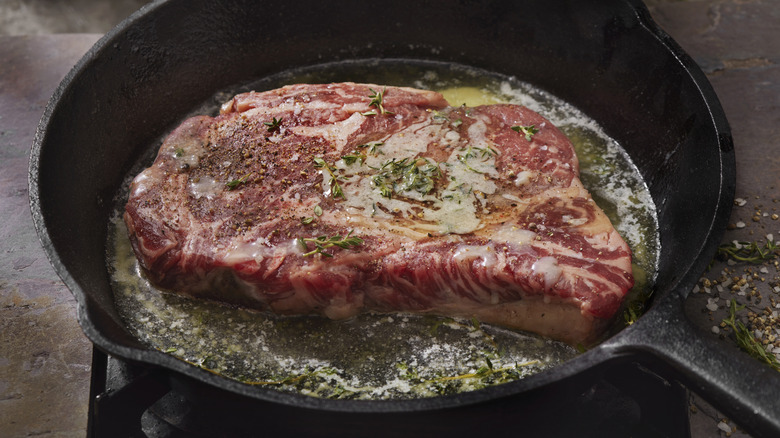 Large ribeye steak cooking with butter and herbs in cast iron skillet