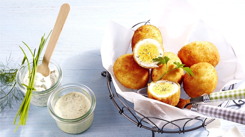 Basket of breaded and fried hard-boiled eggs next to two jars of white sauce