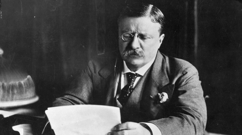 Teddy Roosevelt reading a document at his desk with a monocle