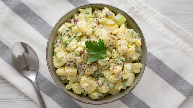 potato salad in a bowl next to a spoon and on a gray and white striped cloth
