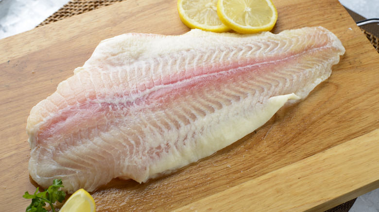 A whole fillet of raw fish sits on a wood cutting board surrounded by slices of lemon