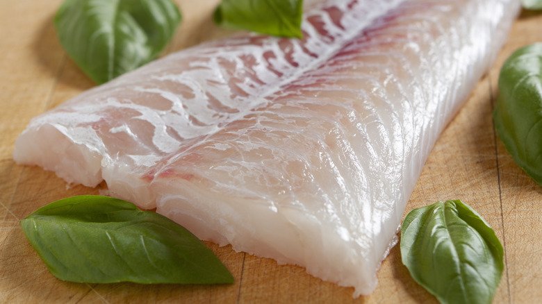 A V-cut of fish sits on a wood cutting board surrounded by springs of basil