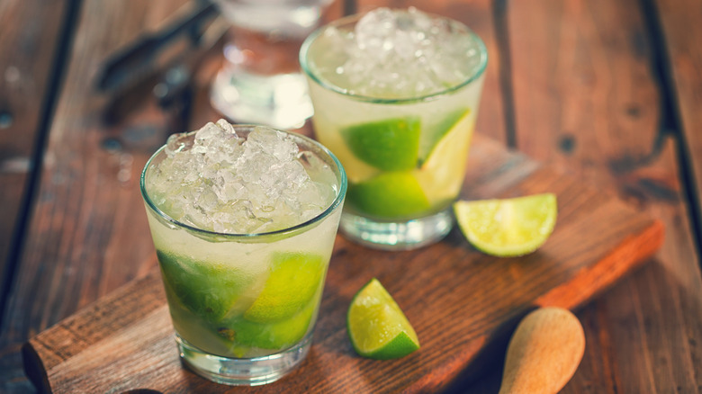 Caipirinha cocktail with crushed ice sits on a wooden table surrounded by limes