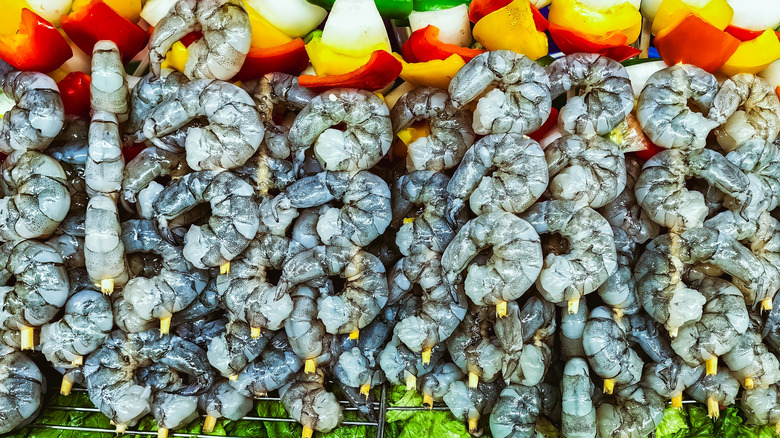 An array of raw shrimp is cooking on skewers on a grill.