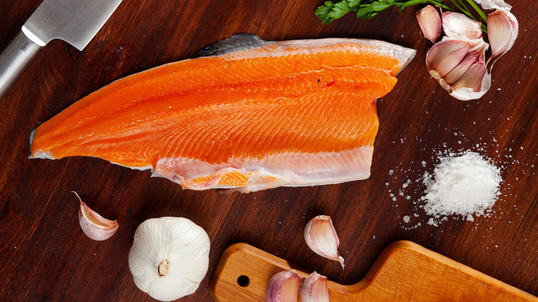 A side of steelhead trout is shown on a wooden counter with garlic, salt, and herbs.