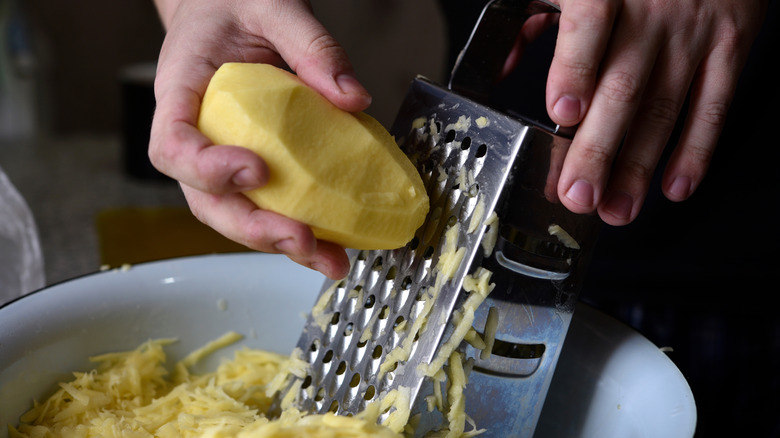 Hands grating a potato into a white bowl using a metal box grater