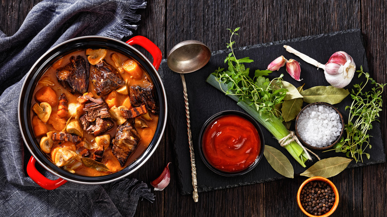 Beef Bourguignon in a pot pot on wooden table with ingredients on the right side atop a cutting board, with a ladle. Denim napkin on left side of the pot.