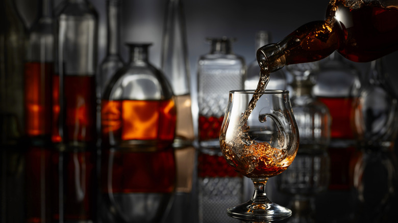 brandy being poured into a snifter on a polished black table and a background of assorted liquor bottles