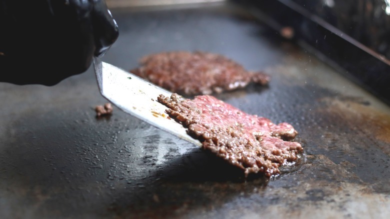 A hand flipping a smash burger with a stainless steel spatula on a flat top grill