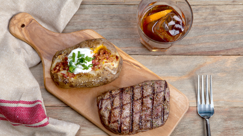 Cooked steak and baked potato on cutting board with a whiskey cocktail