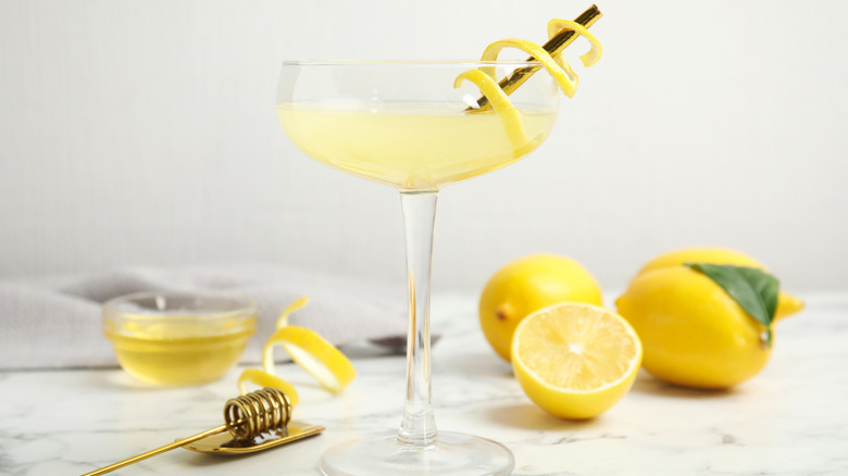 Pale yellow drink in a coupe cocktail glass with sliced lemons and a brass honey dipper