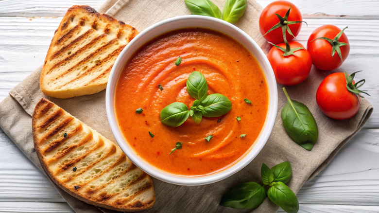 A bowl of tomato soup sits next to two pieces of bread along with tomatoes and basil.