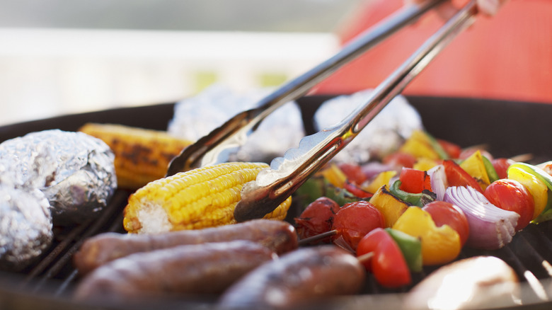 Metal tongs reach for corn on the cob on a grill filled with other vegetables