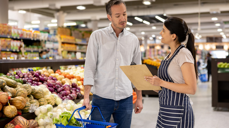 A man holding a shopping basket speaks with a supermarket employee holding a clipboard in the produce section