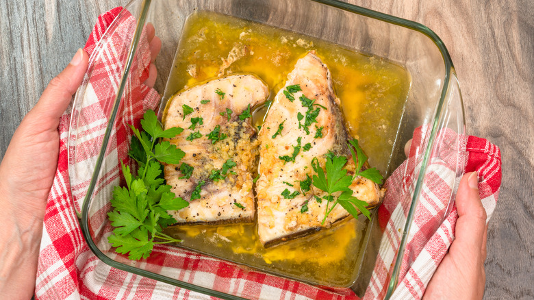 Two filets of white fish sitting in a marinade with herbs in a glass dish