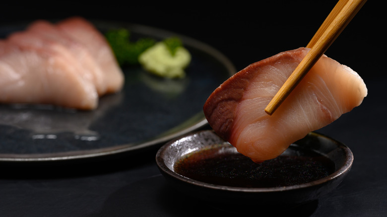 Piece of yellowtail sashimi being dipped into soy sauce with chopsticks