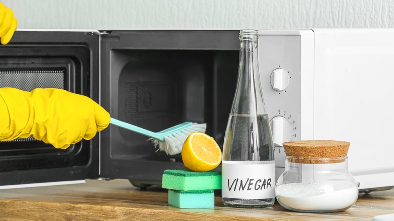 A person wearing rubber gloves using a brush to clean a microwave, beside it sits sponges, half a lemon, a bottle labeled 'vinegar' and a jar of baking soda