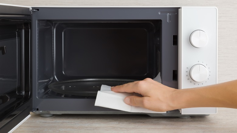 A young woman wiping the inside of an open microwave with a white paper towel