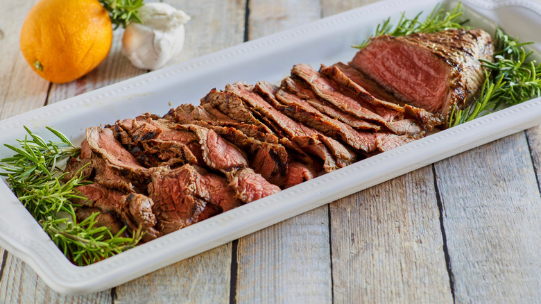 London broil steak sits sliced very thin in a white platter on a light wooden surface