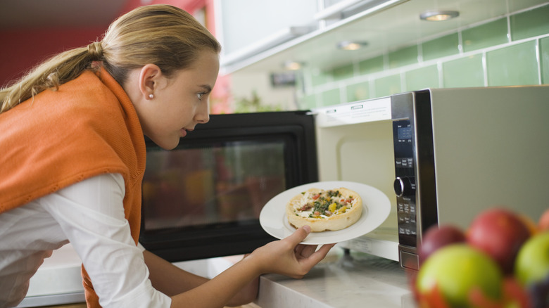 Young woman placing plate with food inside microwave