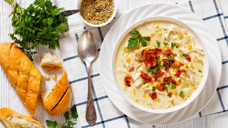 A white bowl of creamy chicken corn chowder sits next to a spoon, herbs, and sliced bread