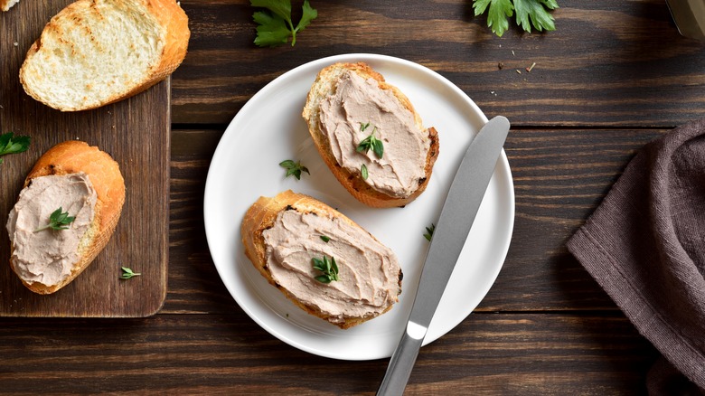 Two slices of bread with chicken liver pate sit on a white plate and slices of bread sit on a cutting board