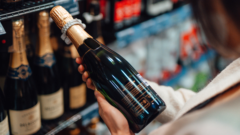 A woman at a grocery store holds a bottle of champagne in the liquor aisle.