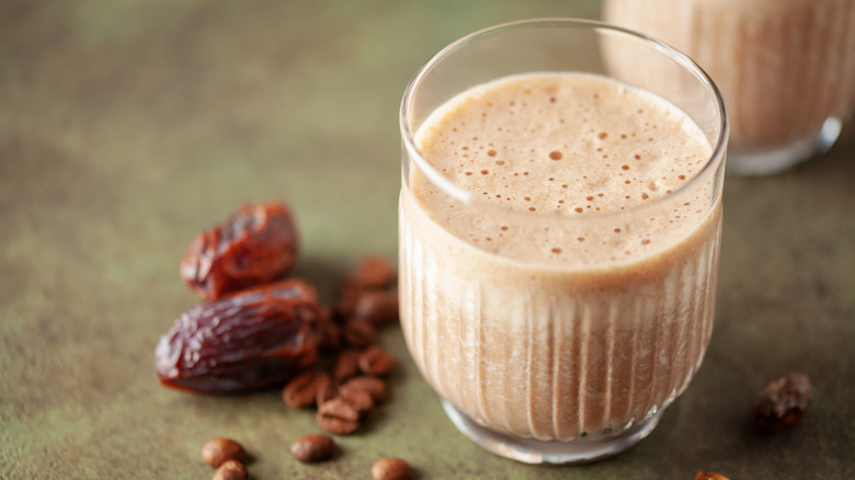 Chocolate, coffee, and date smoothie in a short glass on a textured table. Dates and coffee beans surrounding the glass, with a second glass blurred in the top right corner.