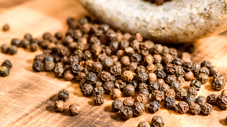 Tellicherry black peppercorns spilling out of a white-gray stone bowl onto a light wooden surface