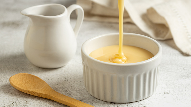 Sweetened condensed milk is drizzled into a white ramekin, with a wooden spoon, and white ceramic cream pitcher