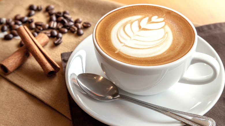 A cappuccino with latte art in a white cup on a saucer with a spoon and coffee beans and cinnamon sticks in the background